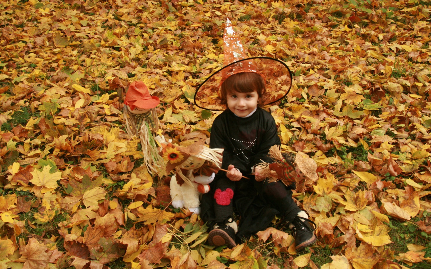 Маленькая девочка в костюме ведьмы сидит на опавшей листве на Хэллоуин