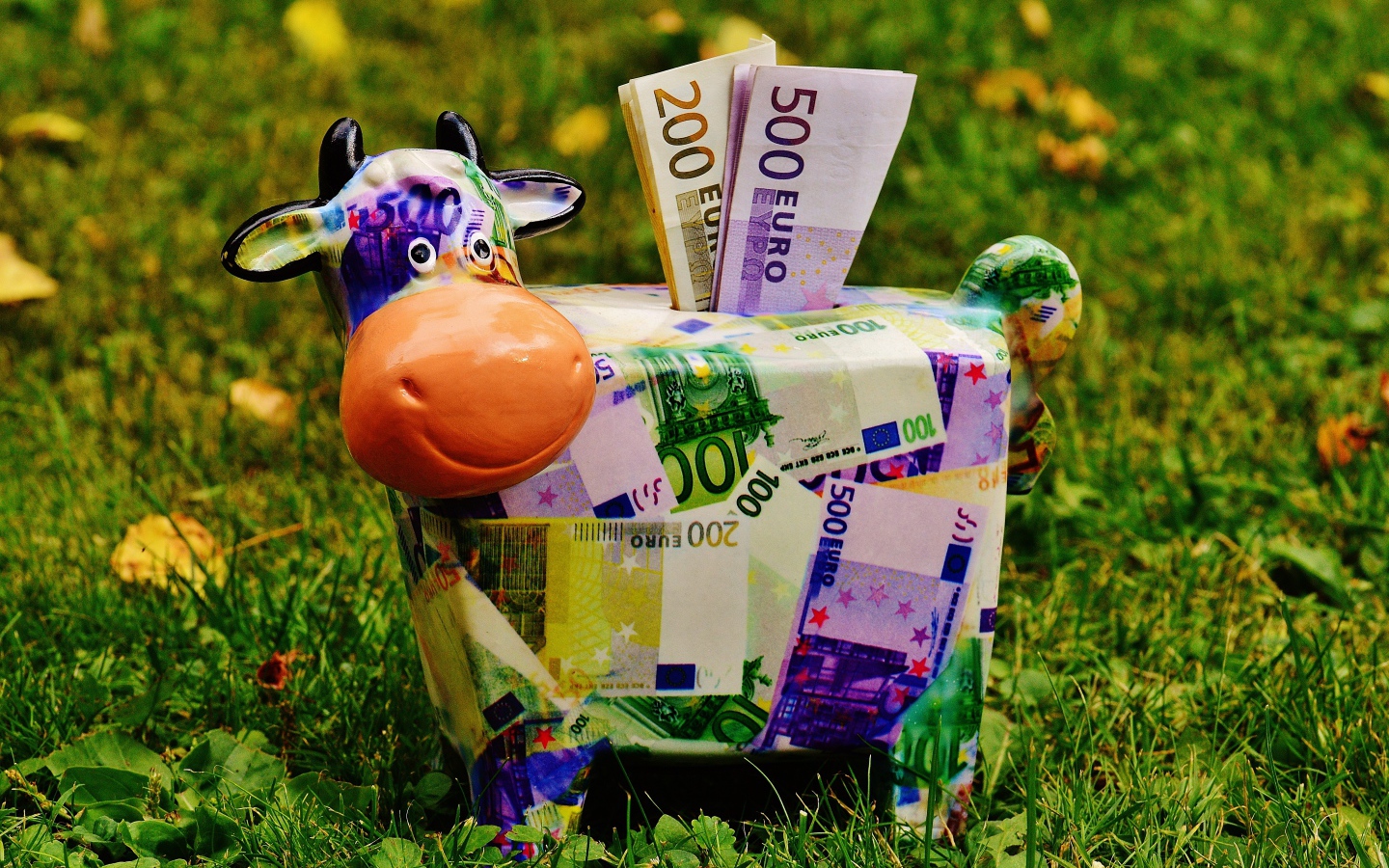 Копилка корова с купюрами евро на зеленой траве 