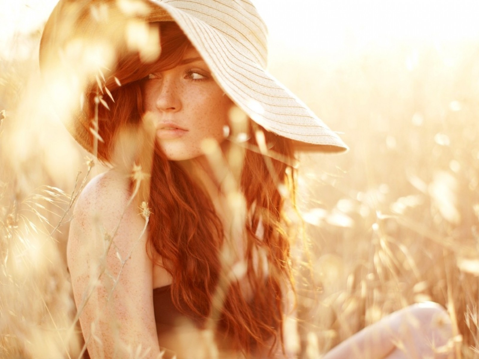 Рыжая девушка в шляпе сидит среди травы на поле