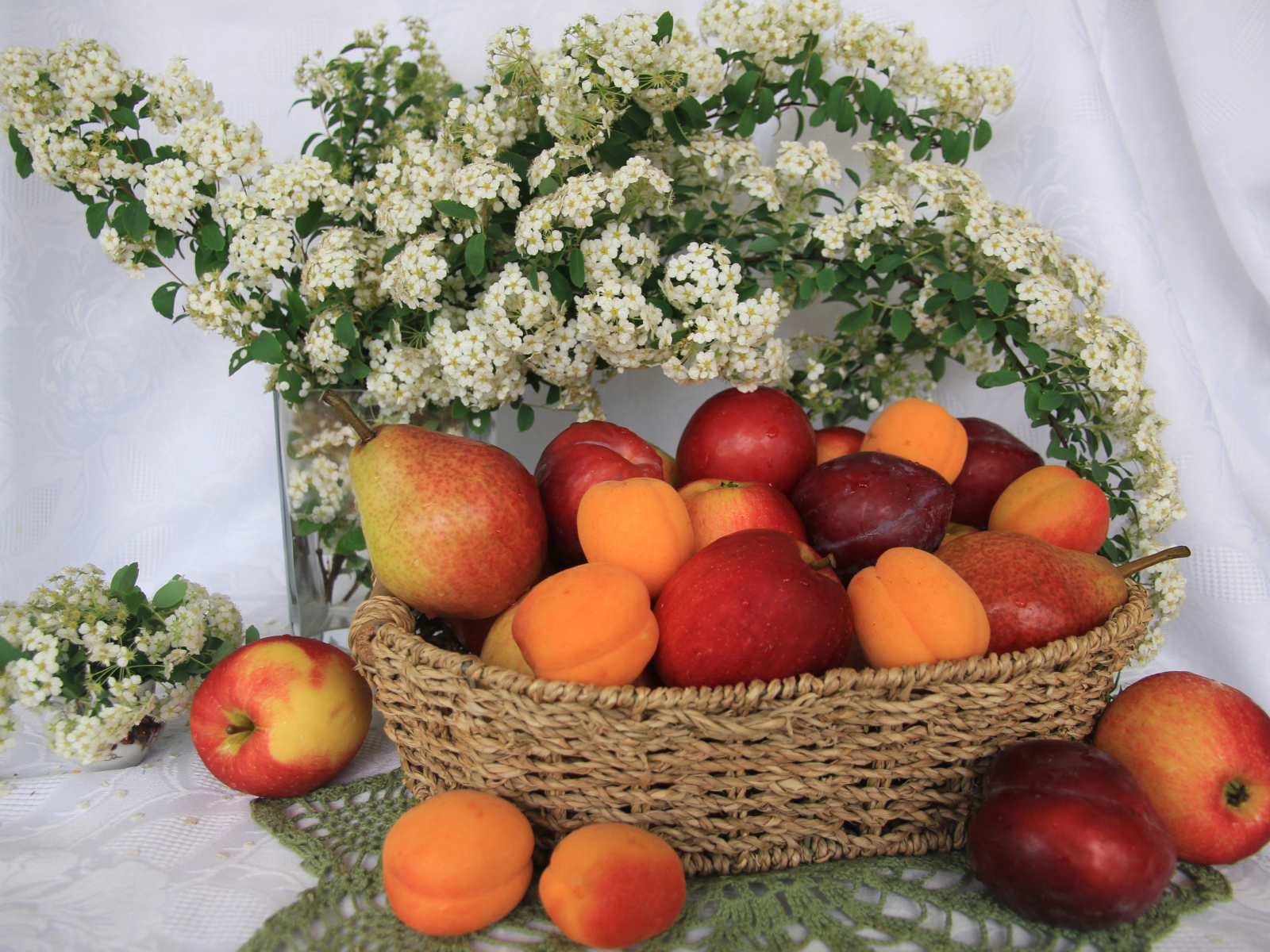 Свежие яблоки, груши, сливы и абрикосы в плетеной корзине на столе с белыми цветами