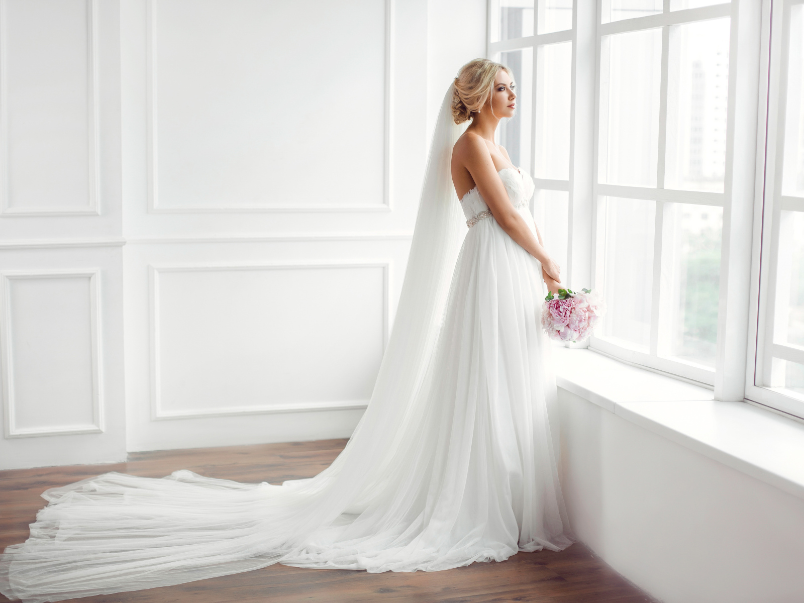 Красивая девушка в белом свадебном платье с букетом стоит у окна