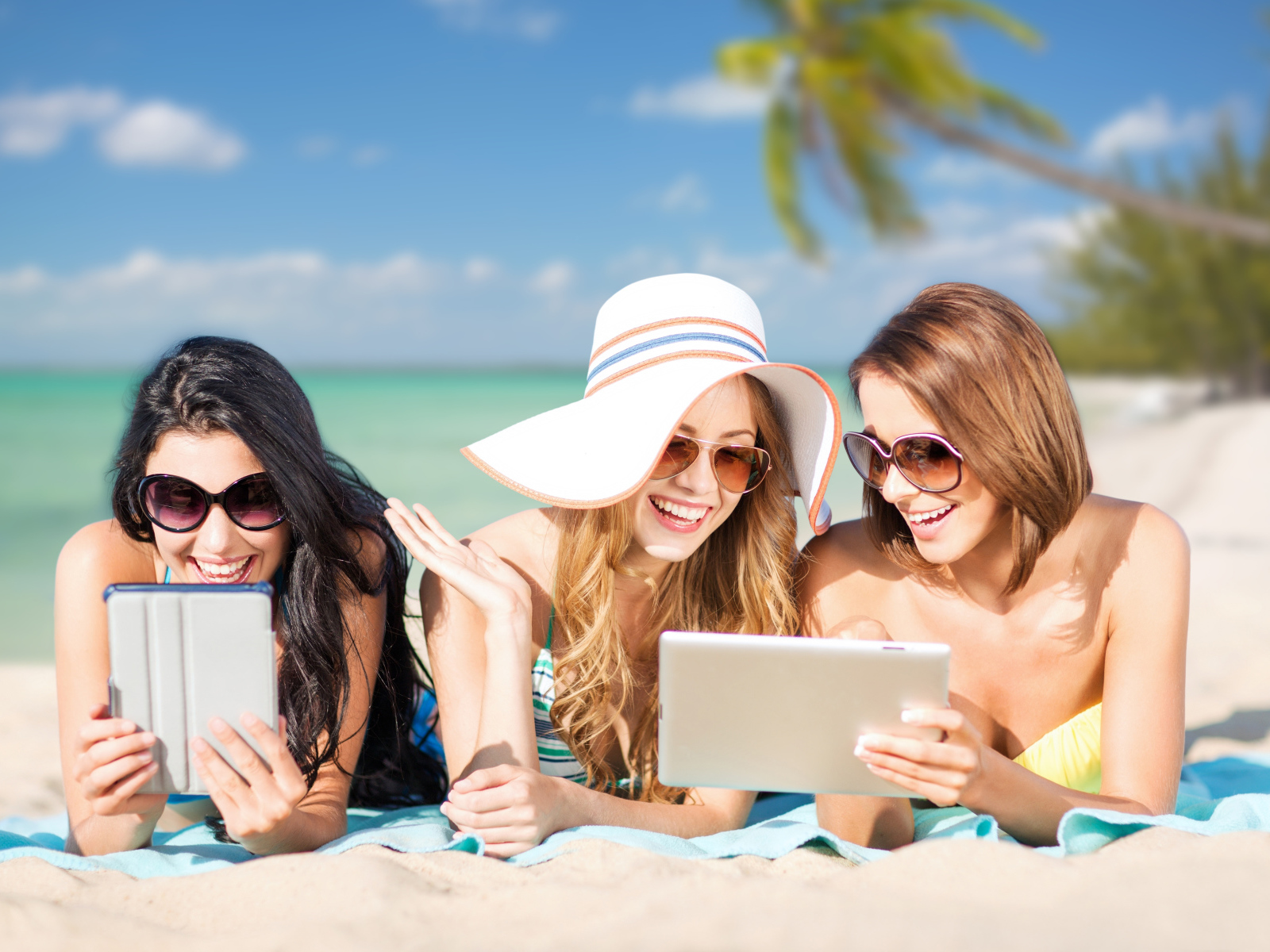 Три улыбающиеся девушки загорают на пляже с планшетами в руках