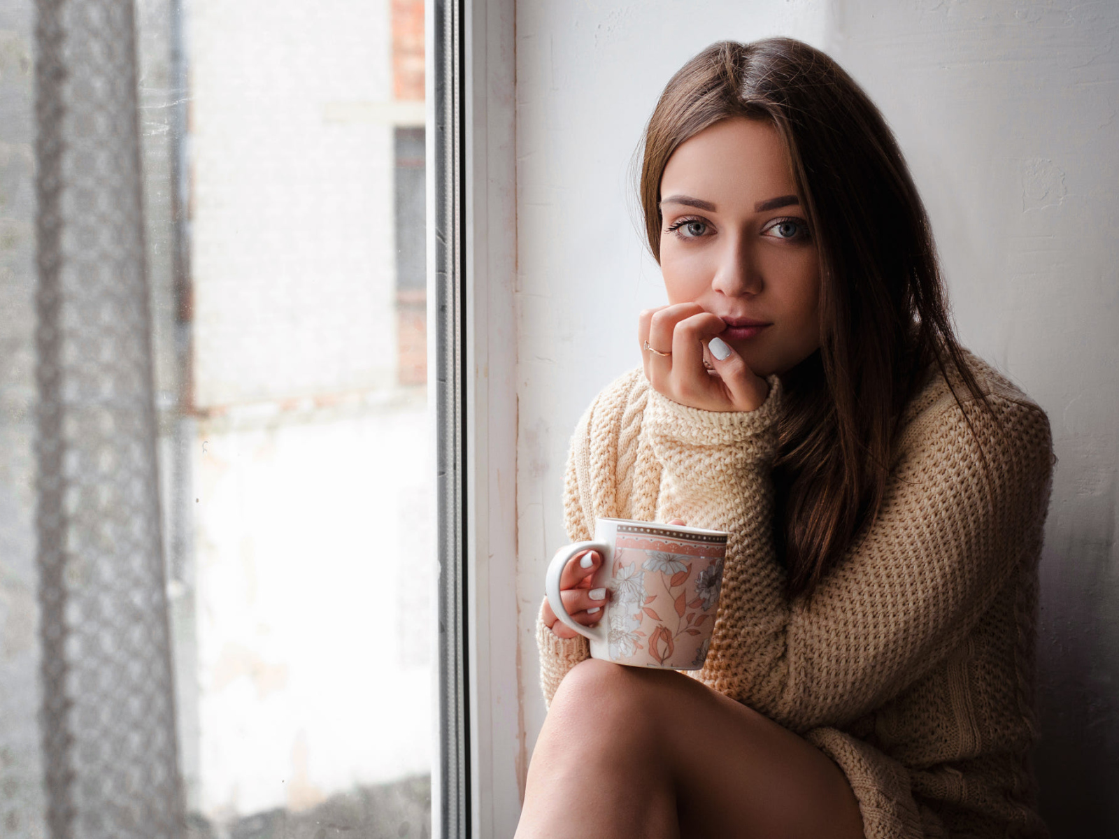 Молодая девушка в свитере сидит с чашкой на подоконнике