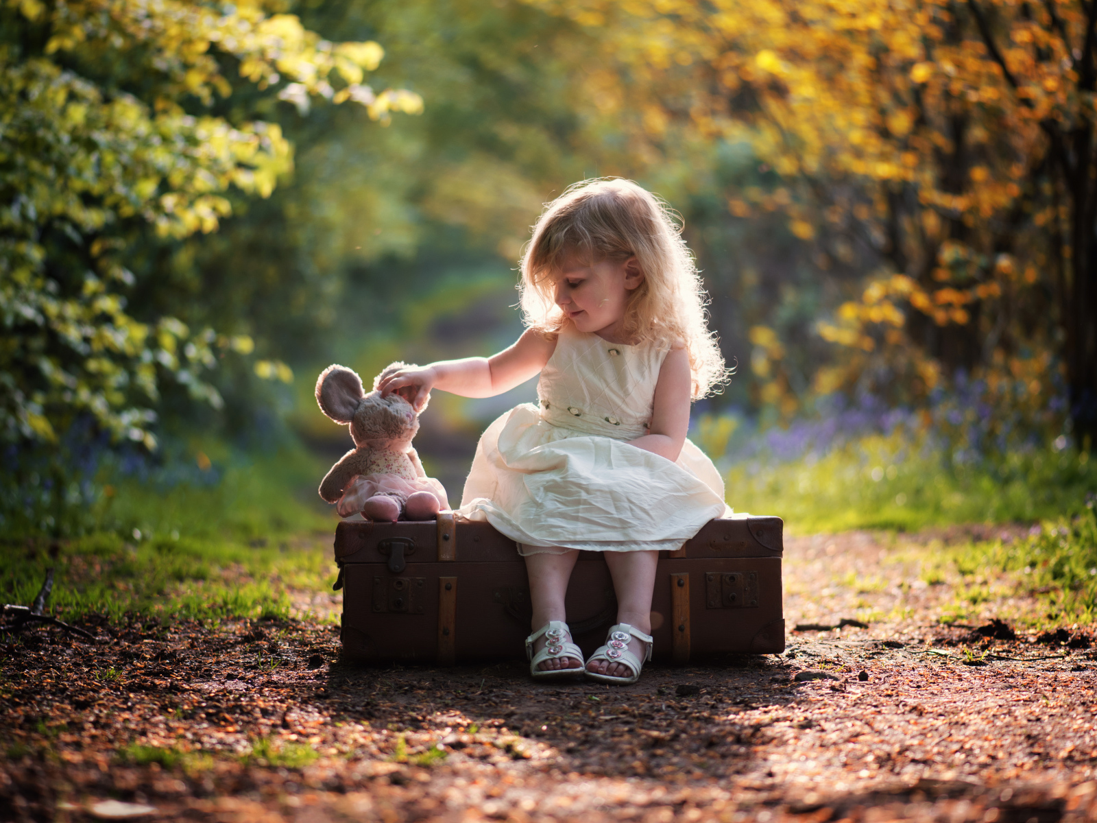 Маленькая девочка в белом платье сидит на большом чемодане с игрушкой