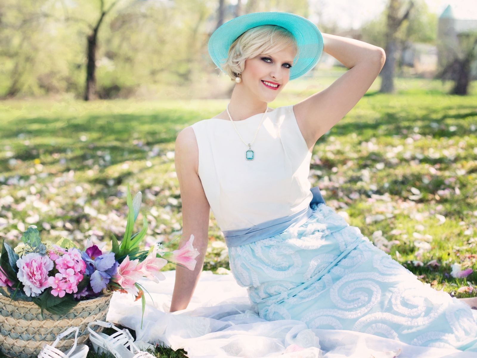 Красивая блондинка в голубой шляпке сидит в парке на траве