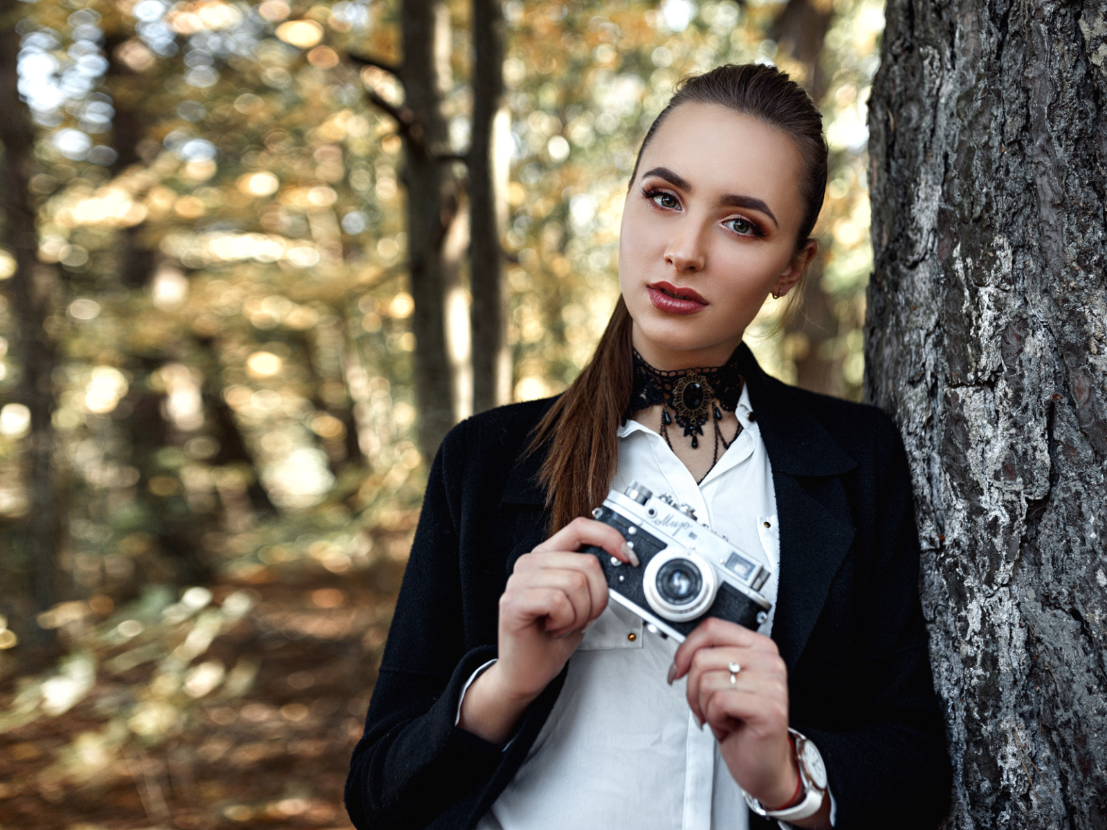 Красивая девушка с фотоаппаратом в руках стоит у дерева