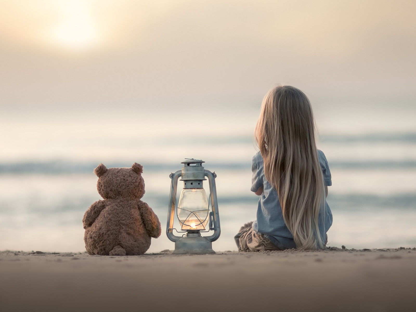 Маленькая девочка сидит на песке с плюшевым мишкой 