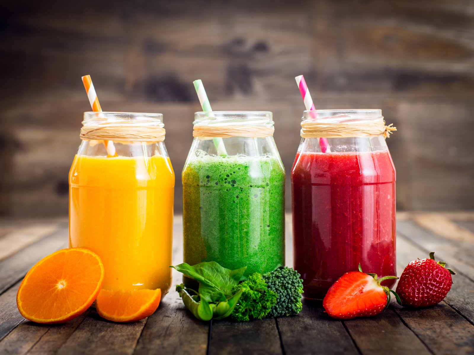 Три банки смузи с трубочкой на столе с апельсином, брокколи и клубникой
