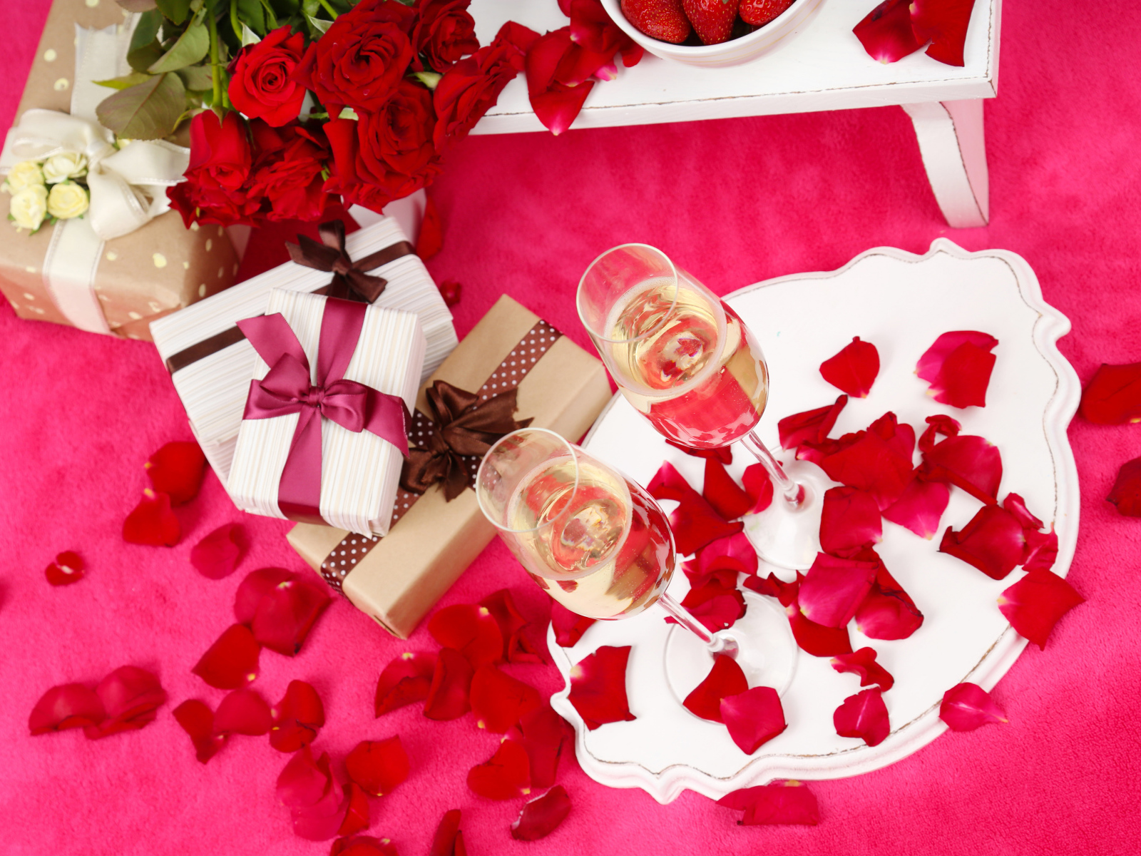 Два фужера с шампанским на красном покрывале с лепестками, подарками и букетом роз для любимой