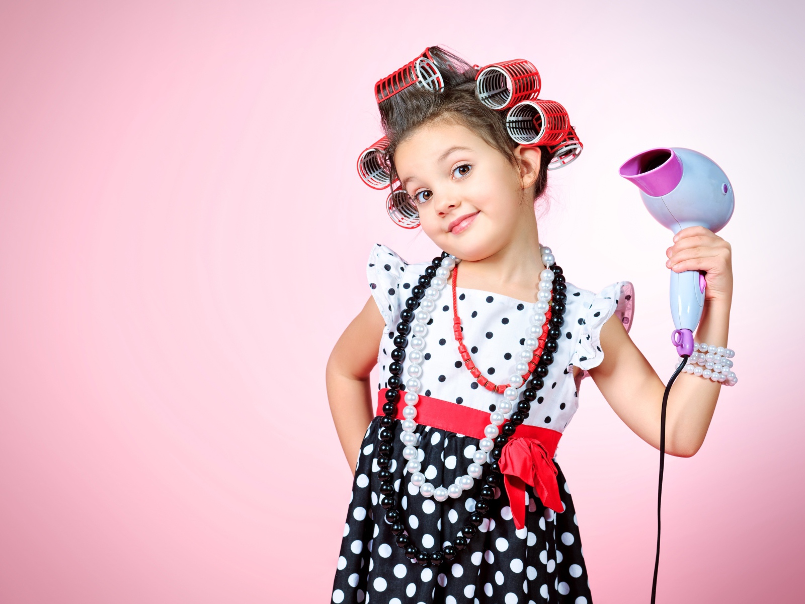 Маленькая девочка с бигудями на голове с феном в руке на розовом фоне