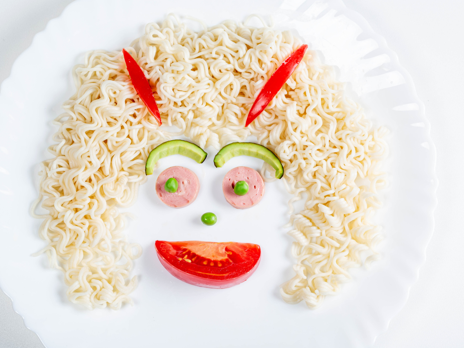 Лицо из вермишели с овощами и колбасой на белой тарелке 