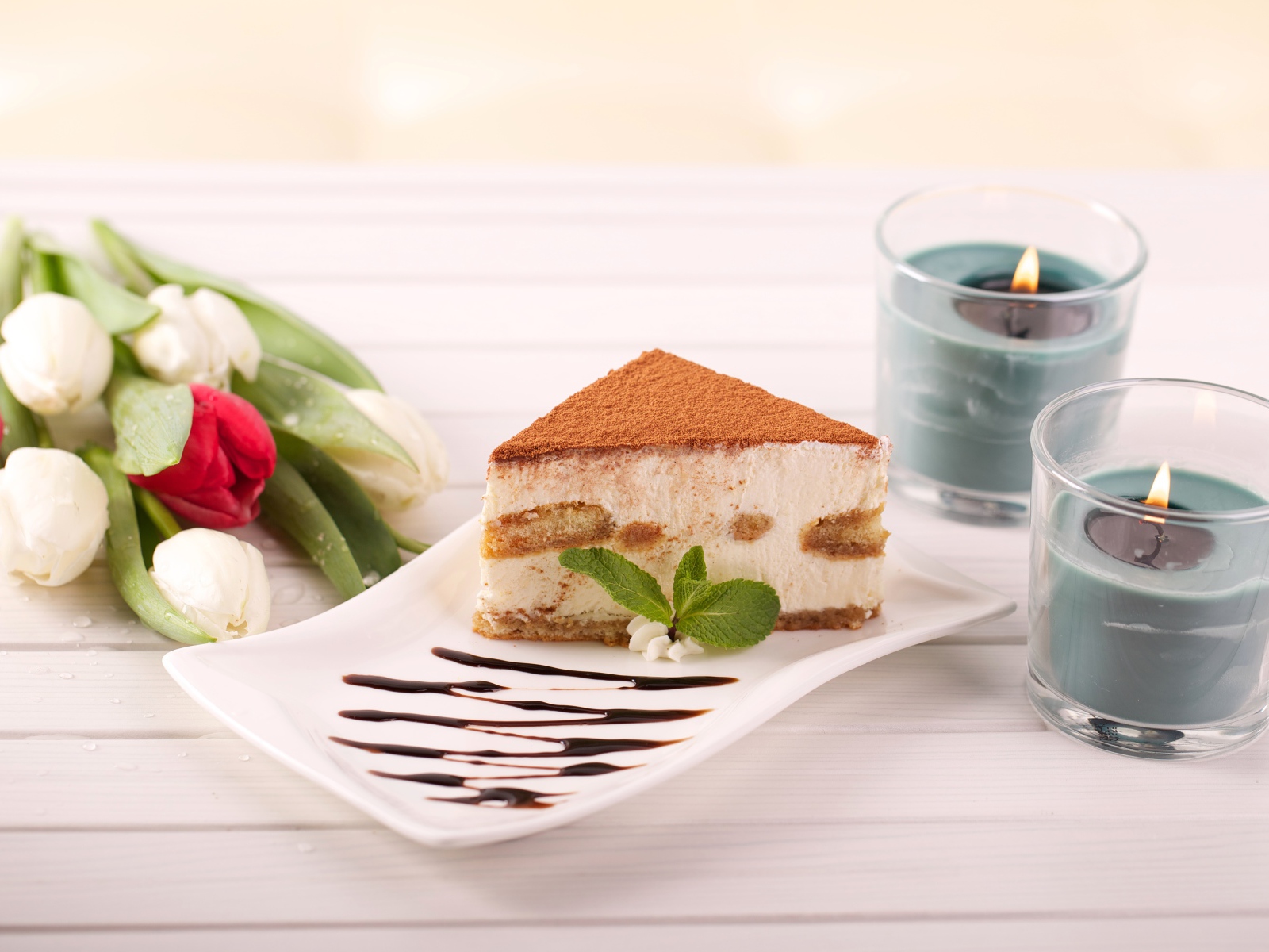 Кусок тирамису на столе с зажженными свечами и букетом тюльпанов