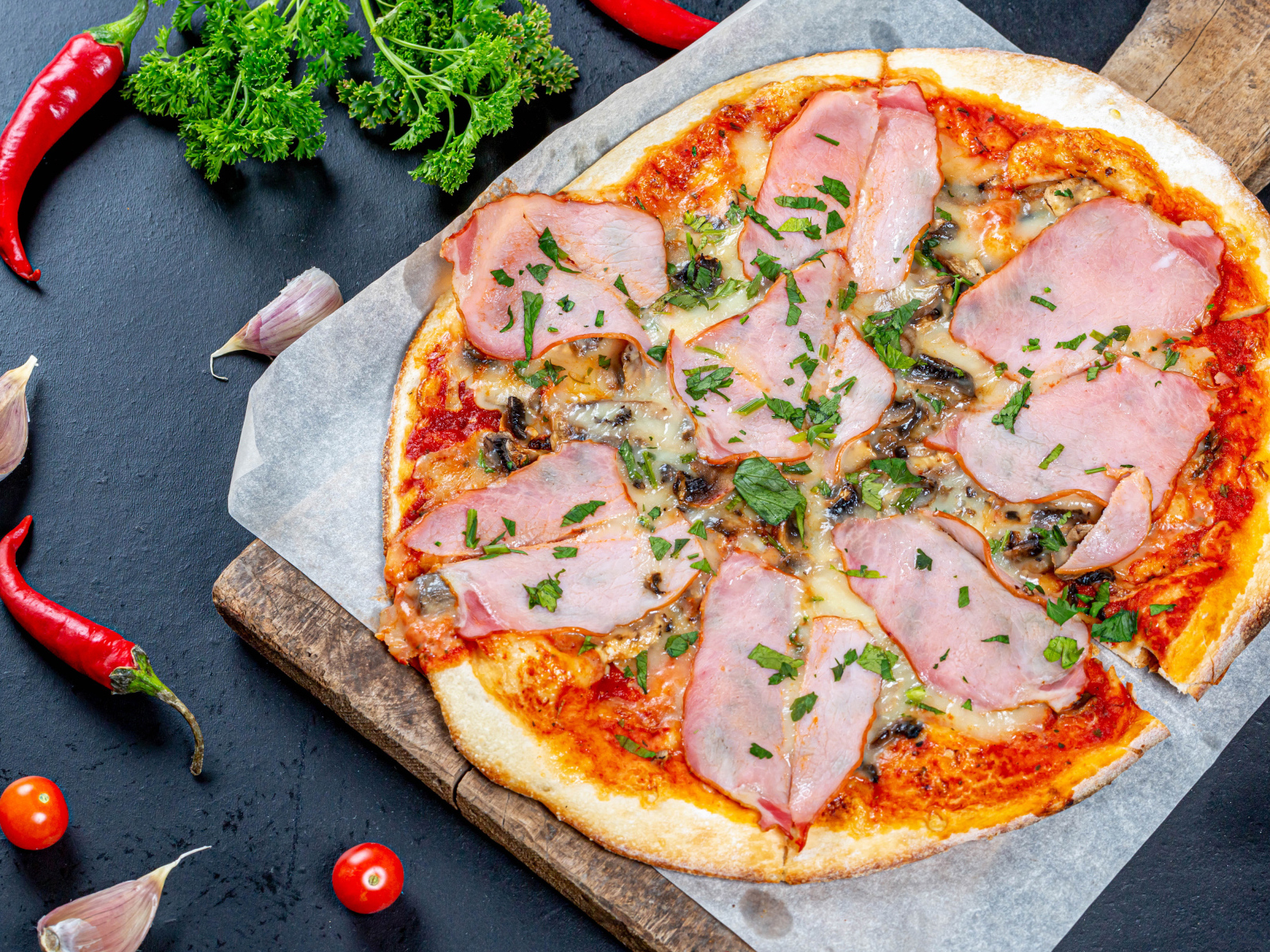 Large pizza with ham on a table with pepper, garlic and parsley