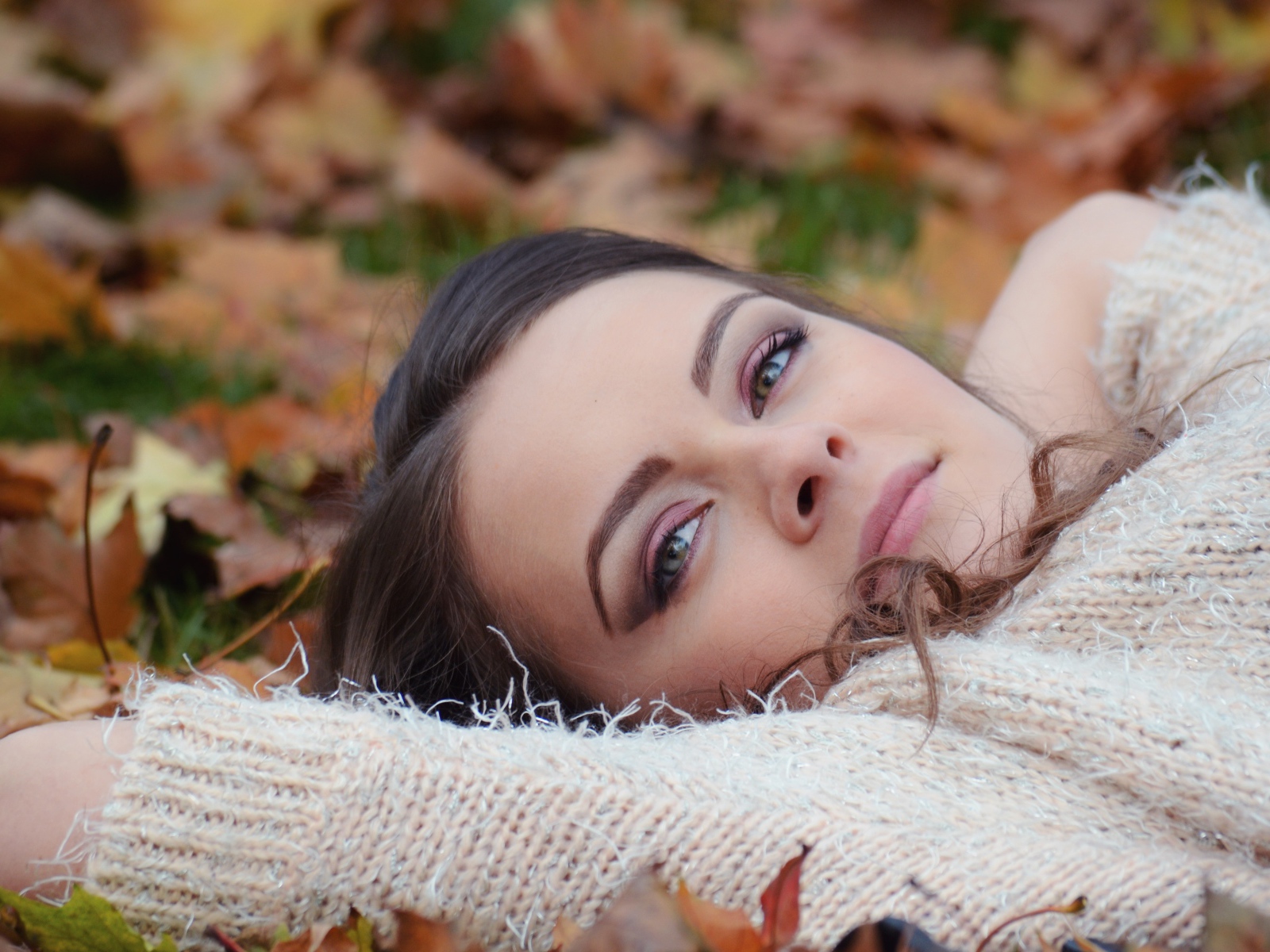 Красивая девушка в теплом свитере лежит на сухой листве 