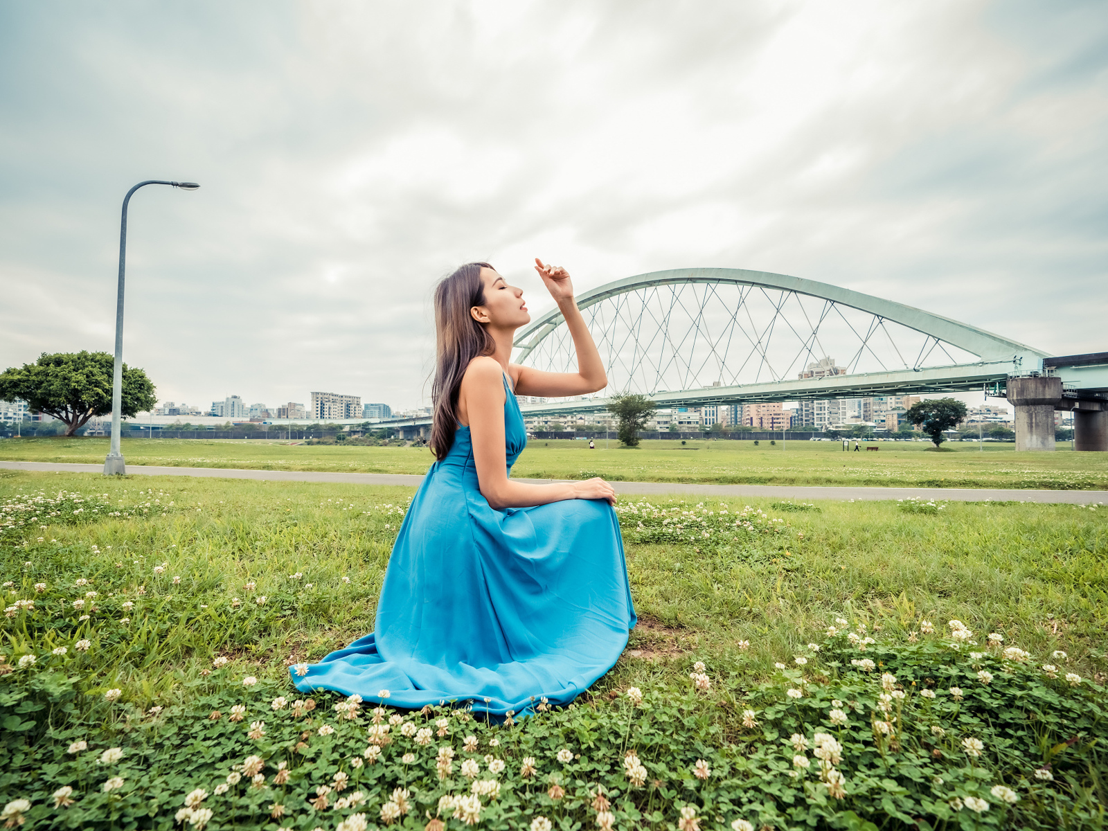 Красивая девушка азиатка в голубом платье  сидит на траве на фоне моста