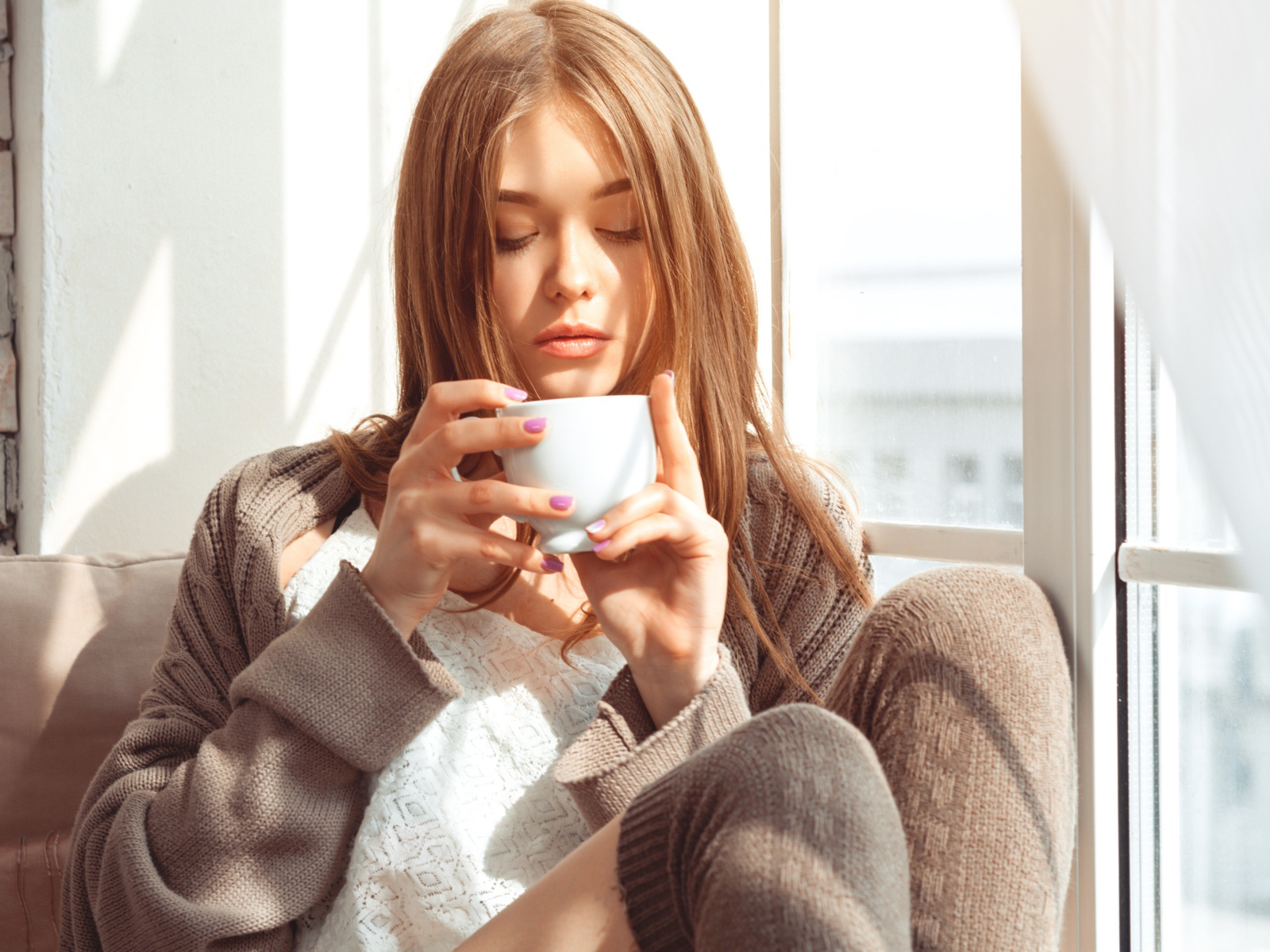 Красивая девушка в свитере пьет кофе у окна