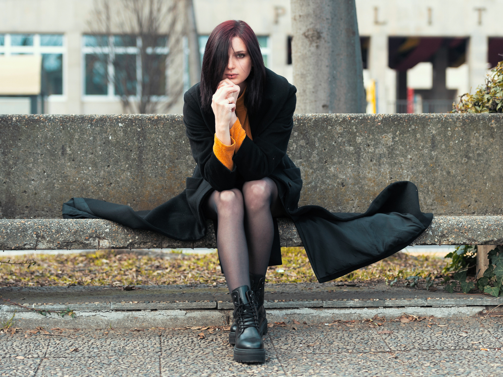 Девушка в черном пальто сидит на бетонной лавке 