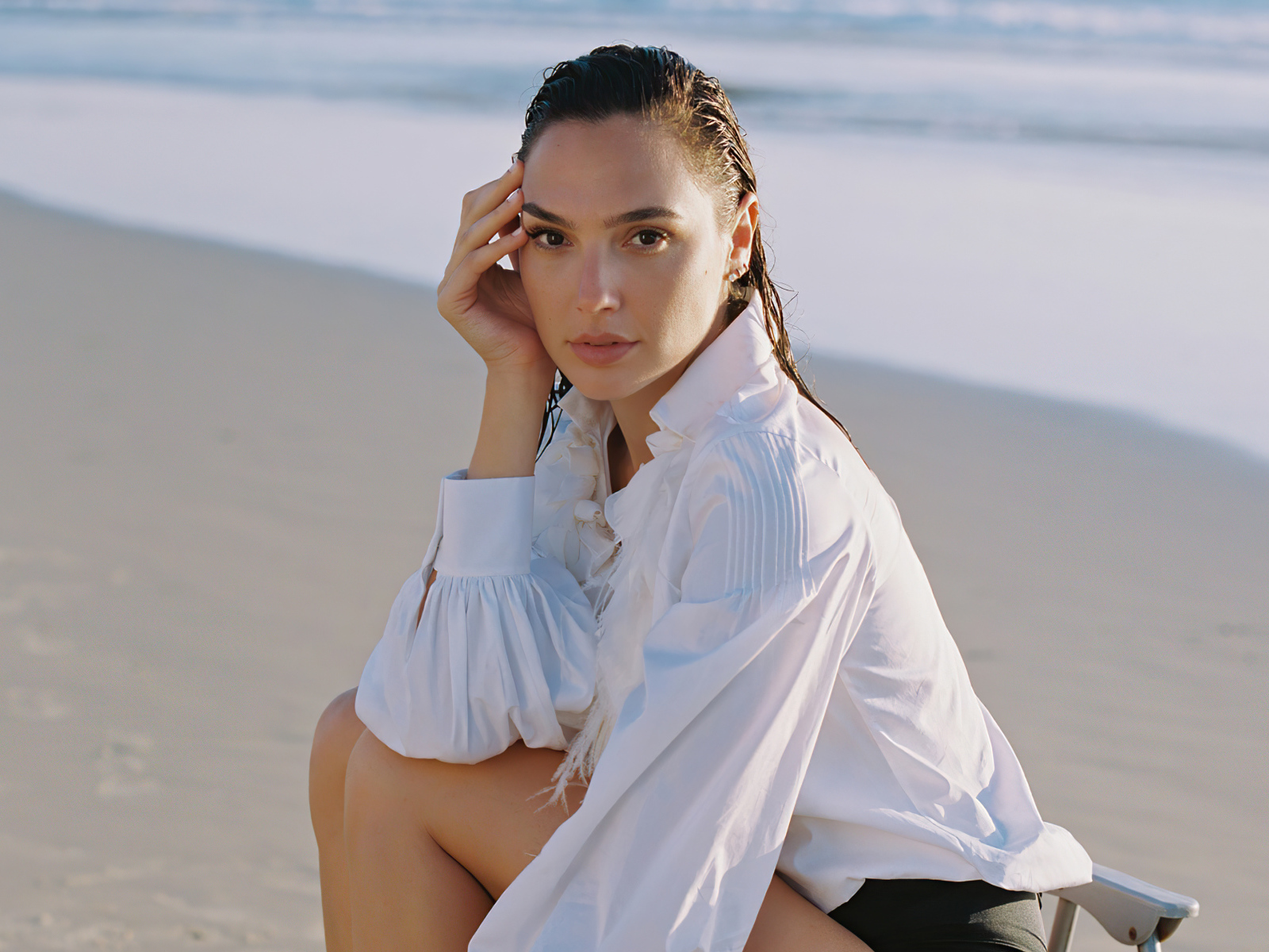 Актриса Галь Гадот в белой рубашке сидит на песке