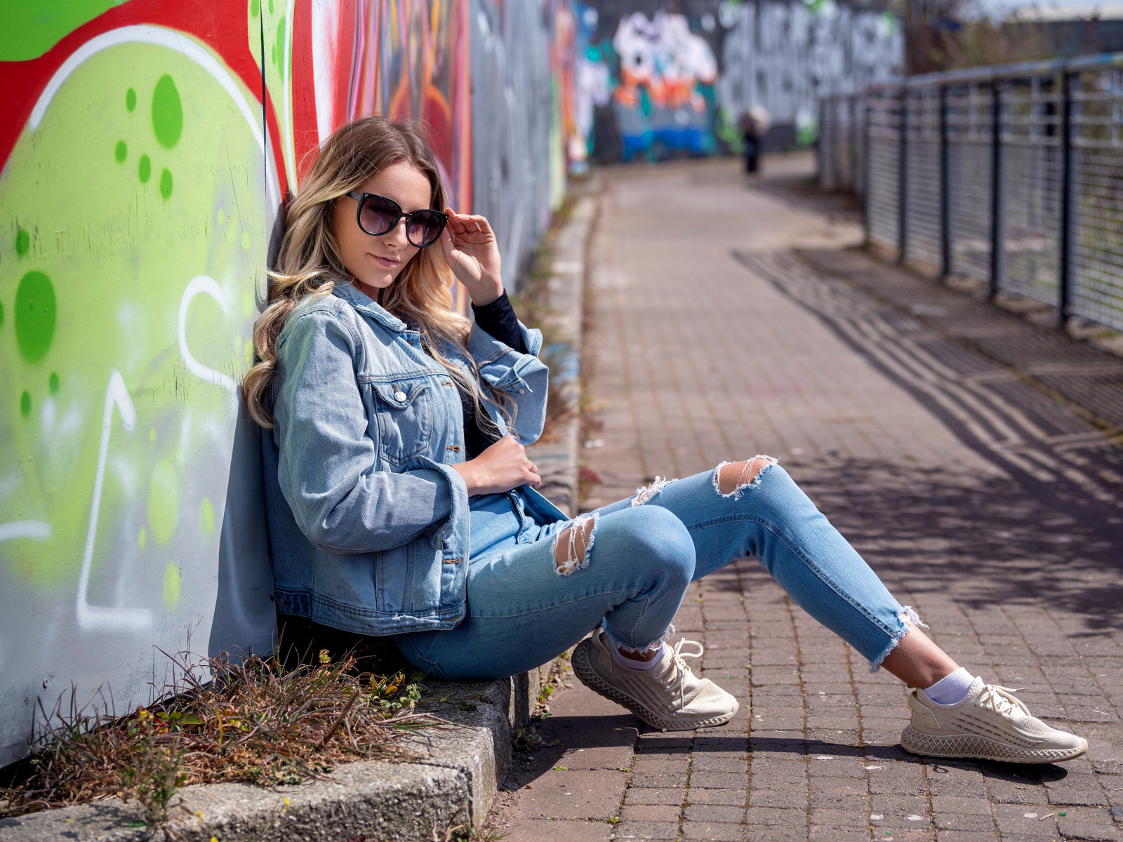 Красивая девушка в джинсовом костюме сидит у стены 