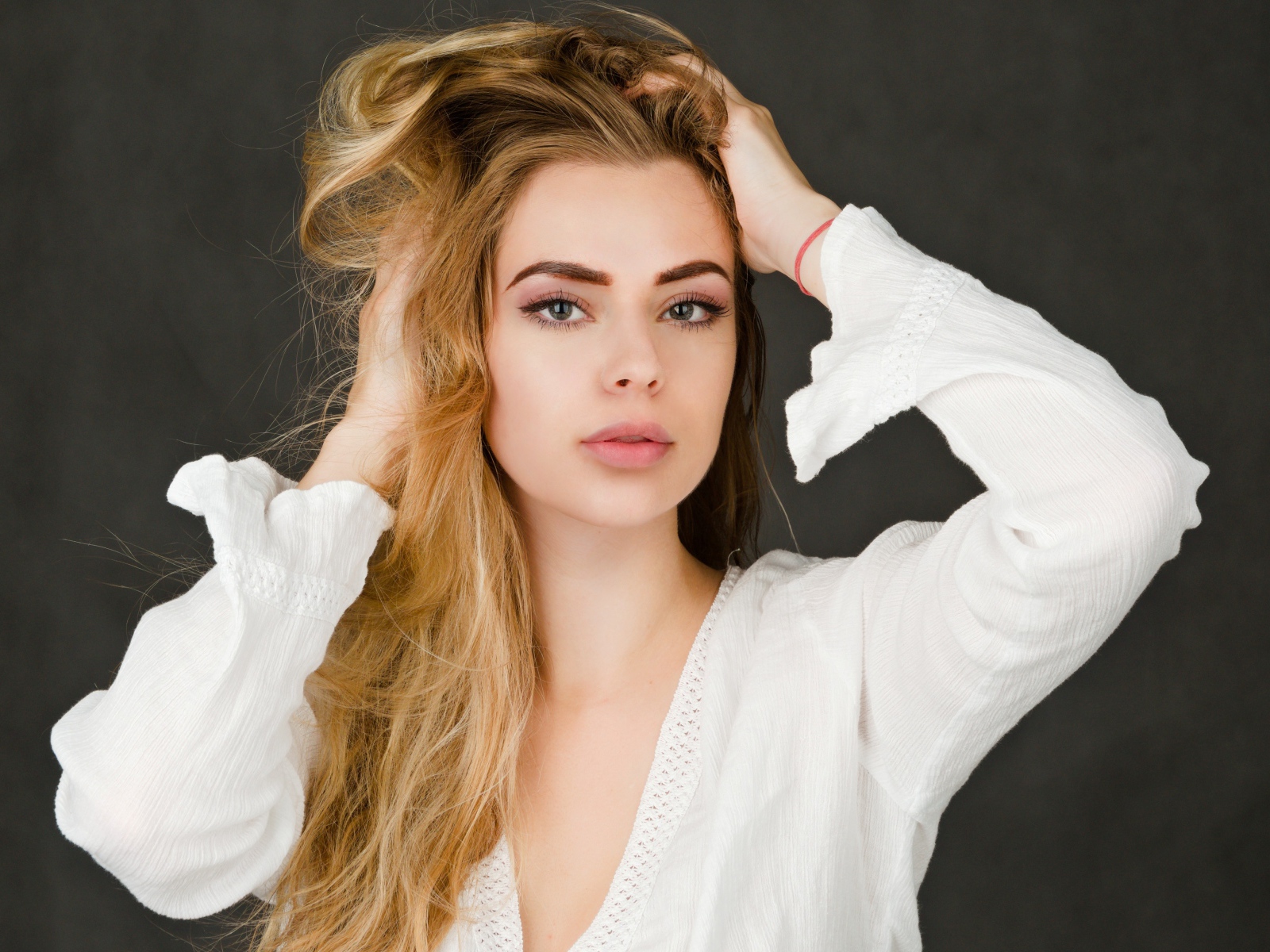 Блондинка в белой кофте с руками в голове на сером фоне 