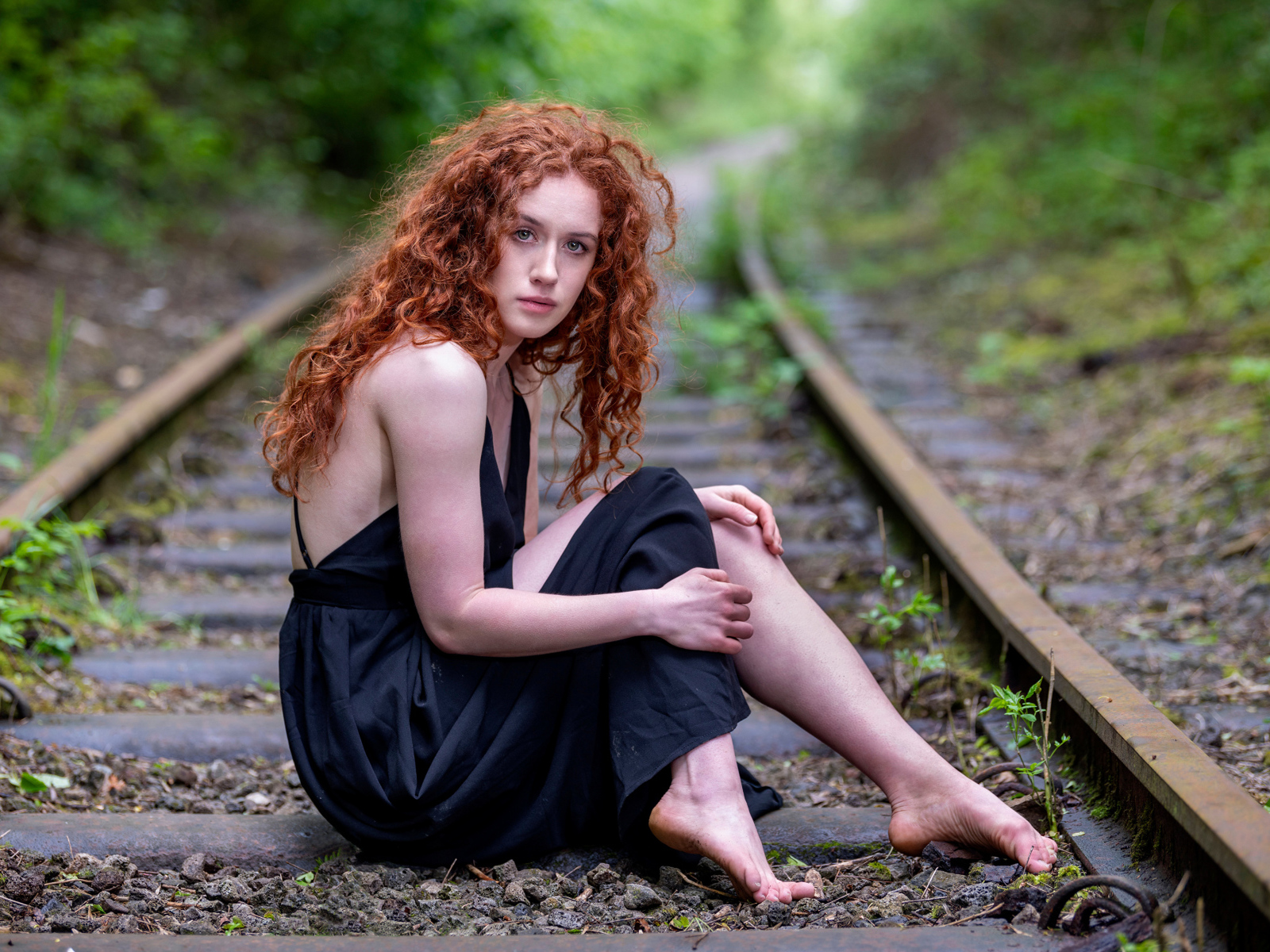 Рыжеволосая девушка в черном платье сидит на путях
