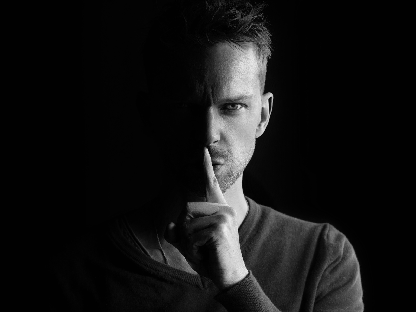 Молодой мужчина закрыл рот пальцем на черном фоне 