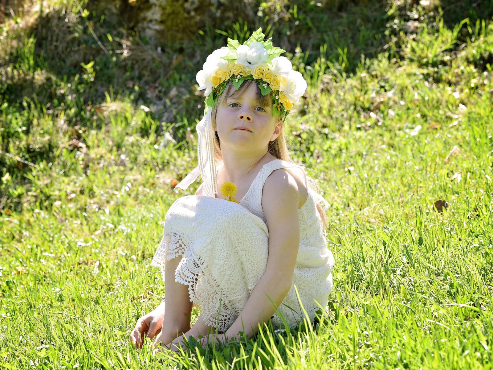 Маленькая девочка с венком на голове сидит на зеленой траве