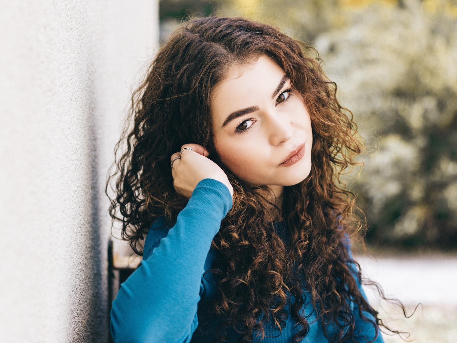 Кареглазая девушка с кудрявыми волосами стоит у стены