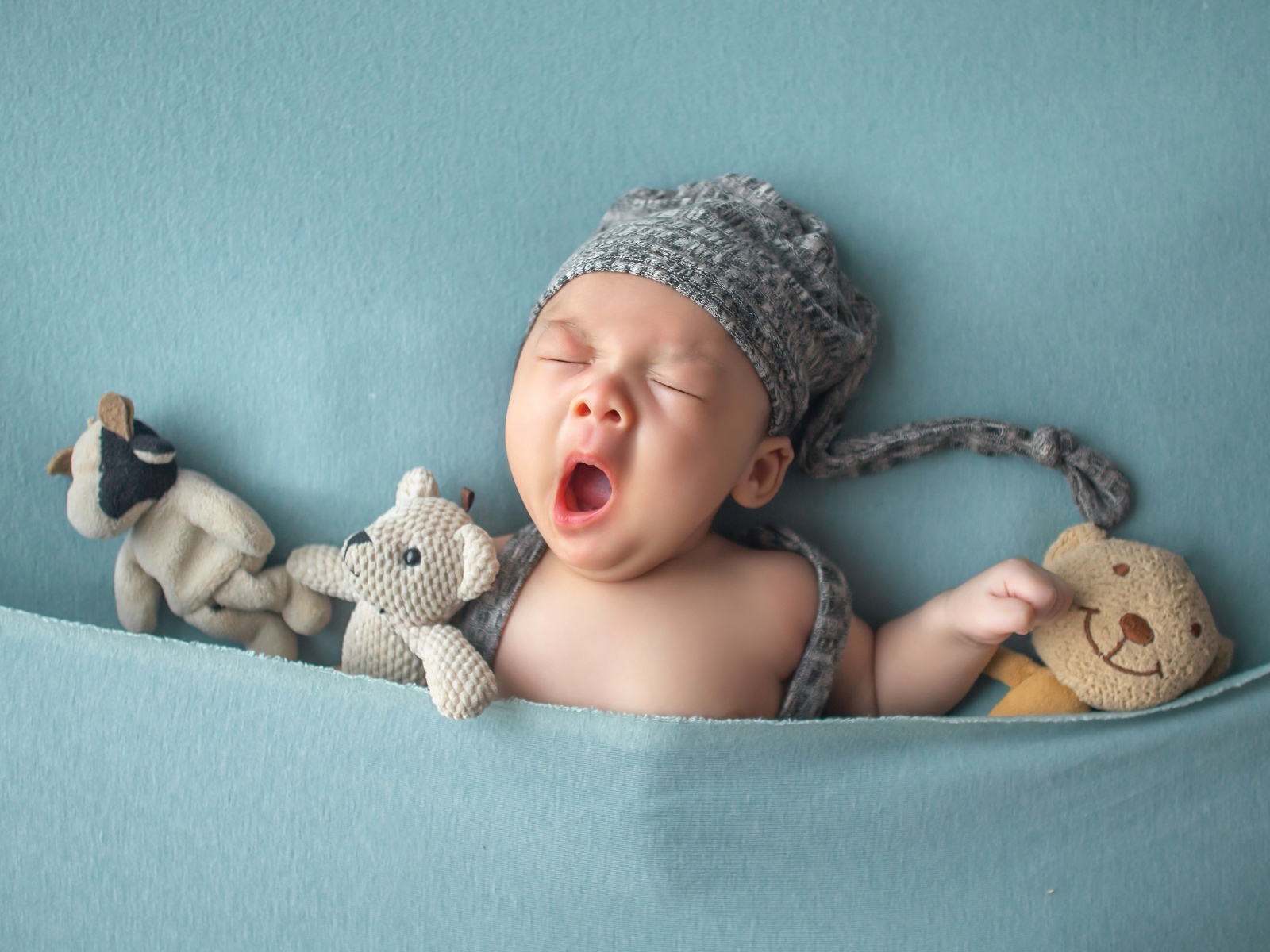 Зевающий малыш с игрушками под серым покрывалом