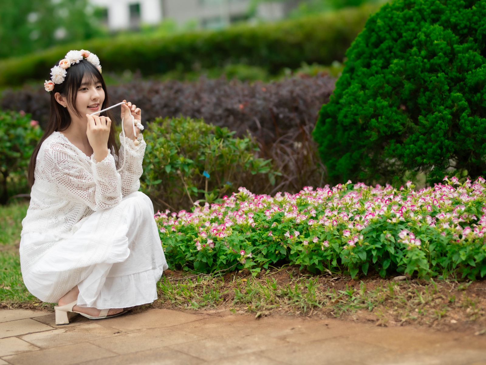 Милая азиатка в белом наряде сидит у клумбы с цветами