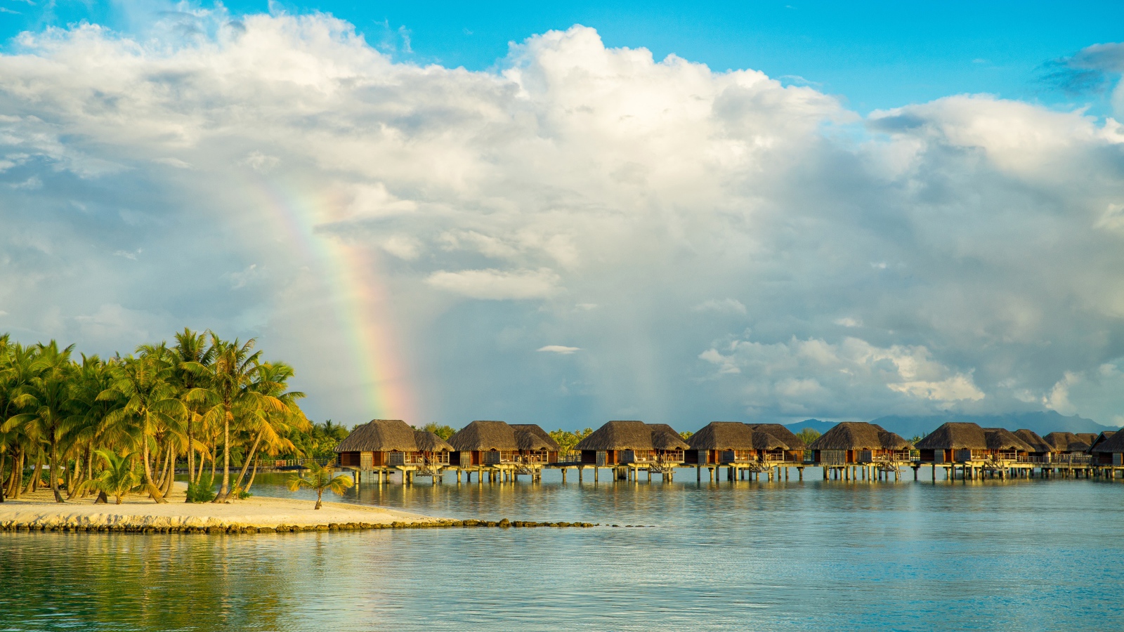 Бунгало в воде на тропическом пляже под облачным небом с радугой