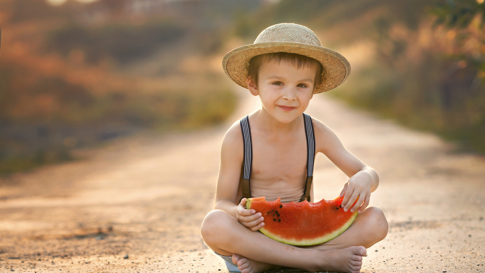 Маленький мальчик в шляпе сидит на дороге с долькой арбуза