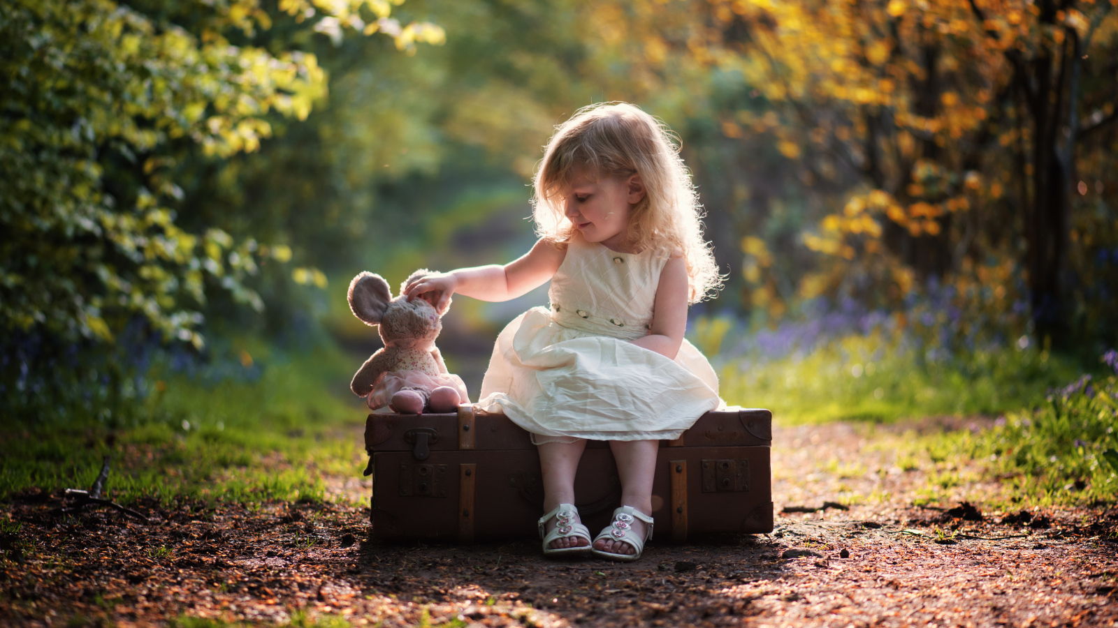 Маленькая девочка в белом платье сидит на большом чемодане с игрушкой