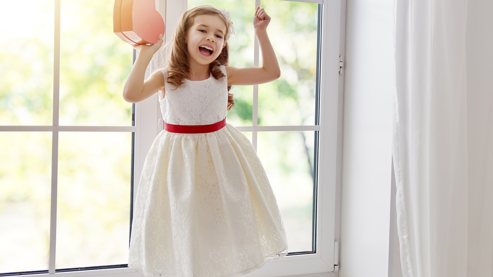 Маленькая девочка с коробкой в форме сердца в руках стоит на подоконнике