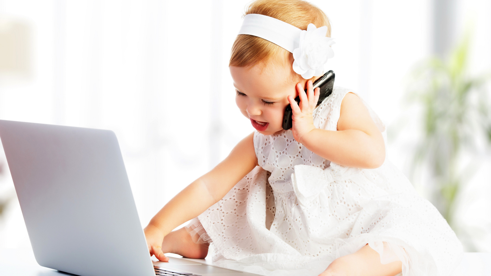 Маленькая девочка в белом платье сидит с телефоном у ноутбука