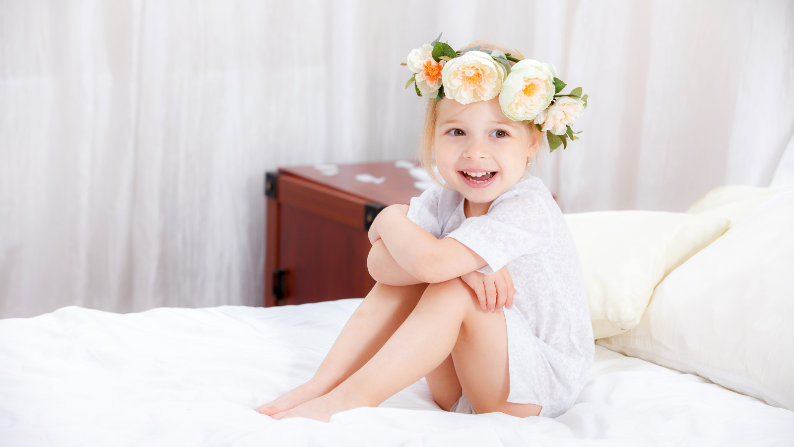 Улыбающаяся девочка с венком на голове сидит на кровати