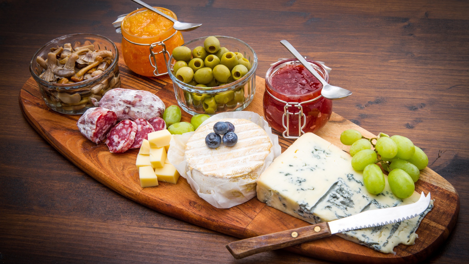 Сыр, грибы, маслины, колбаса, виноград и джем на разделочной доске 