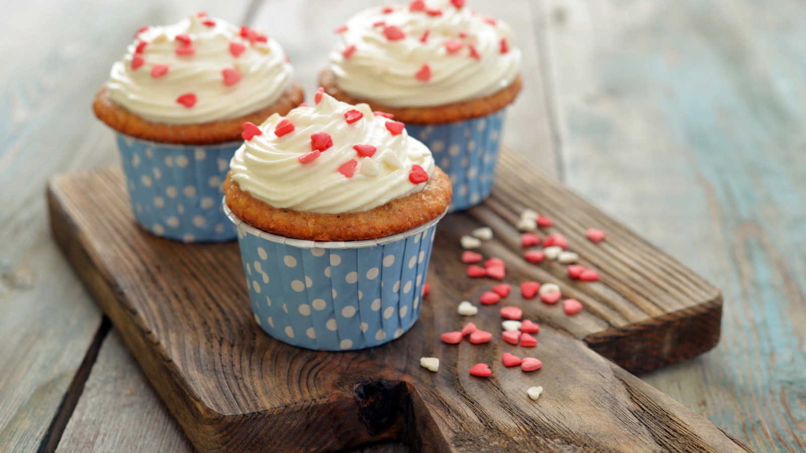 Три кекса со сливками украшены сердечками стоят на разделочной доске