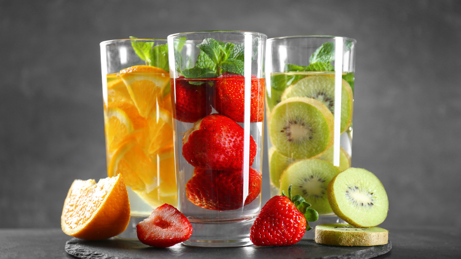 Напитки в стаканах с клубникой, киви и апельсинами на сером фоне
