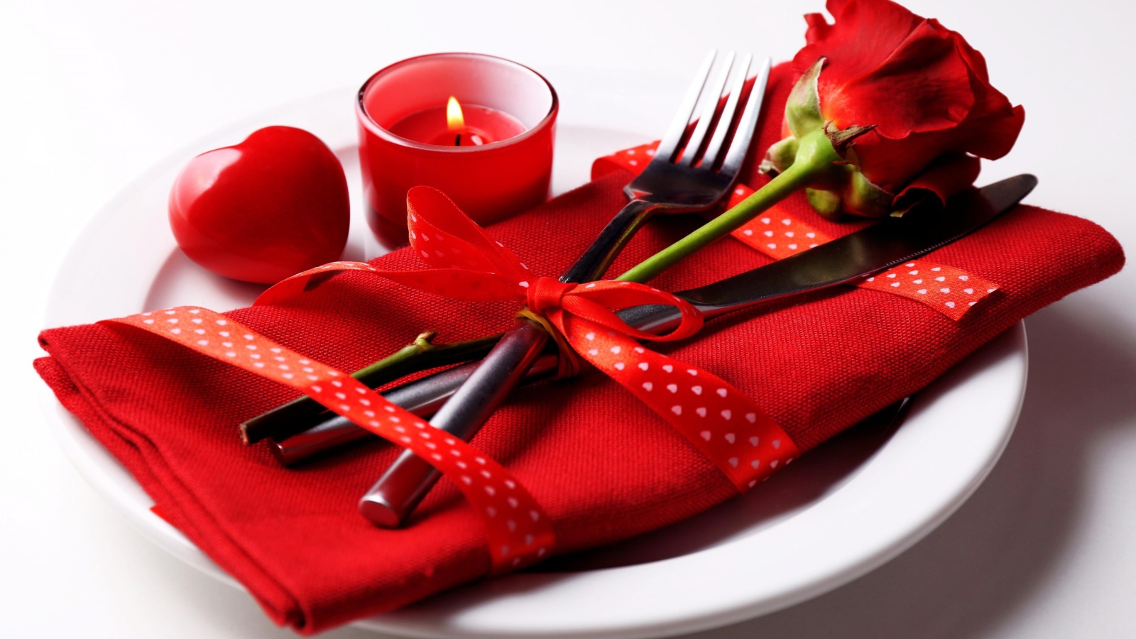 Роза, столовые приборы и зажженная свеча на белой тарелке 