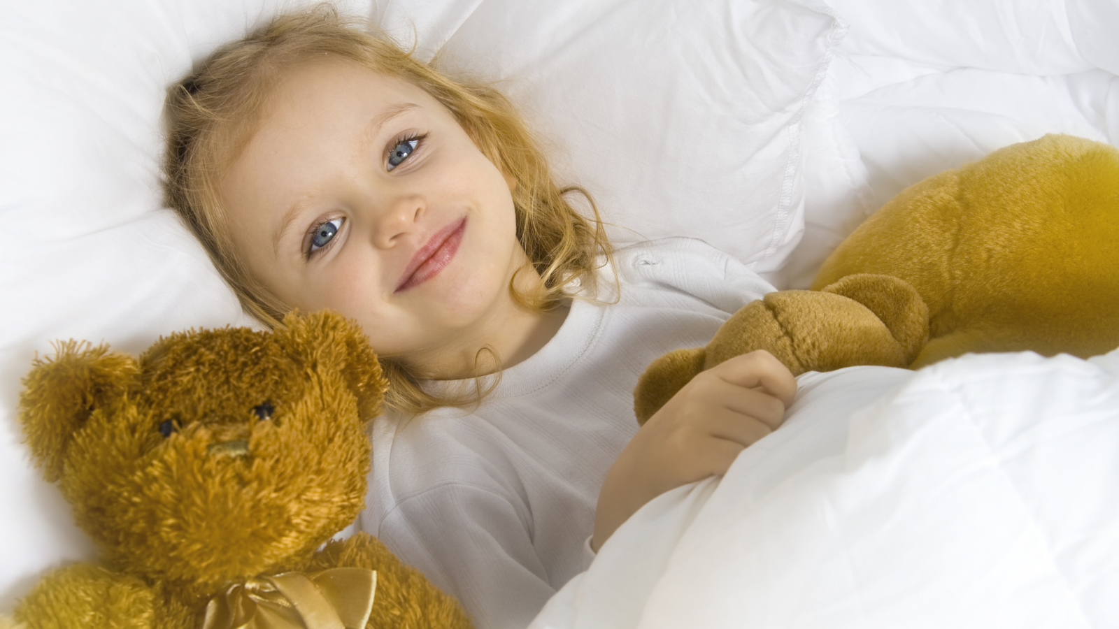 Милая голубоглазая девочка с мягкими игрушками лежит под одеялом