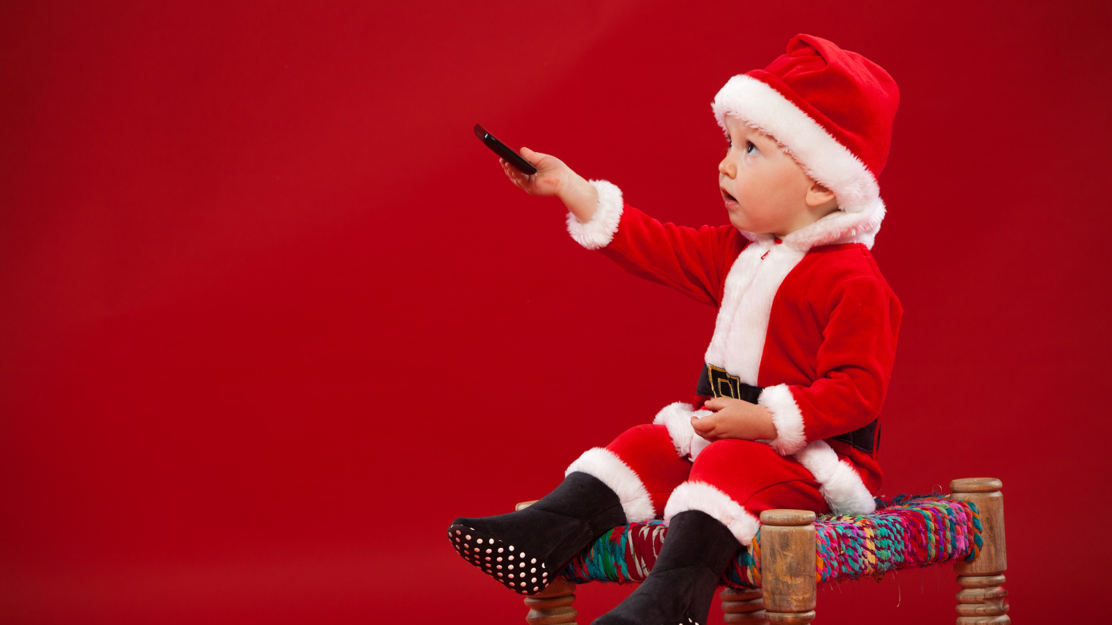 Маленький мальчик в костюме Санта Клауса сидит на стуле на красном фоне