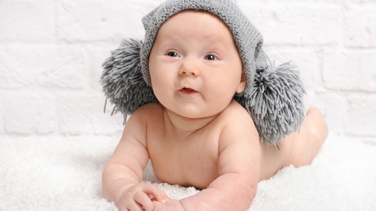Улыбающийся грудной ребенок в серой шапке с бубонами