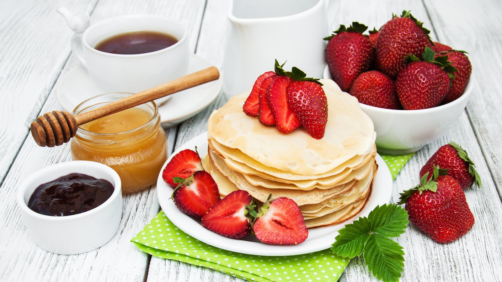 Тонкие блины на столе с ягодами клубники, медом, чаем и джемом