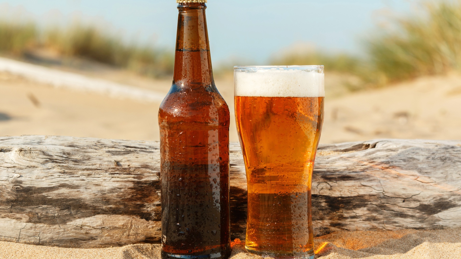 Бутылка пива и бокал стоят на песке 