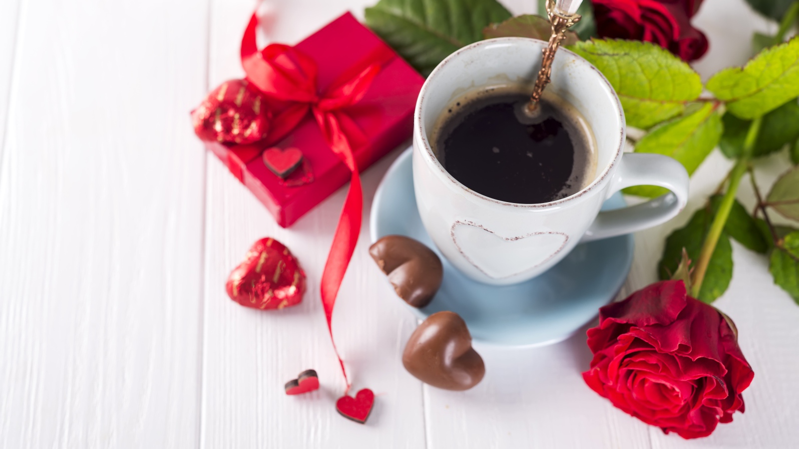 Чашка кофе на столе с шоколадными конфетами, подарком и красной розой