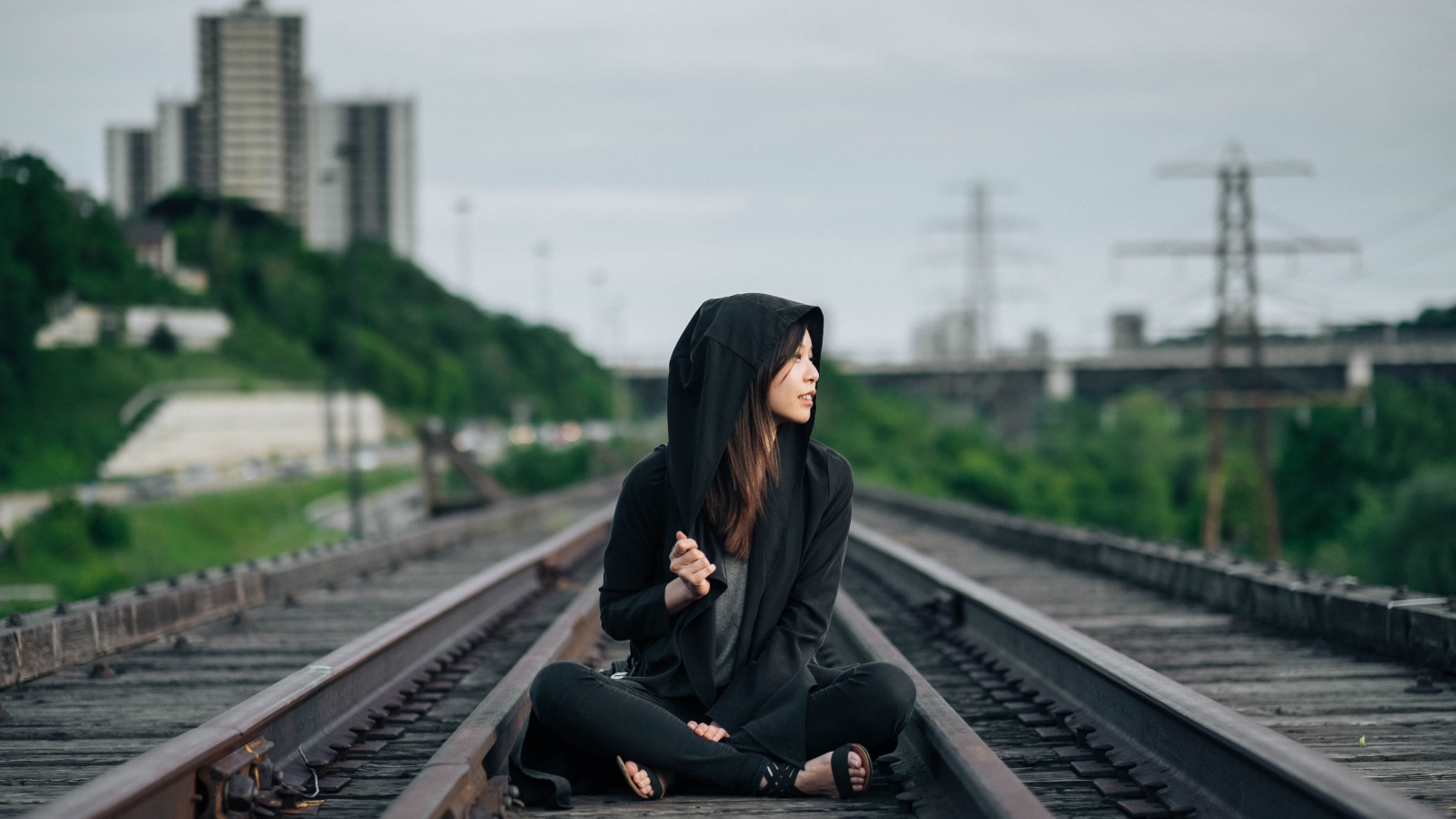 Девушка азиатка в черном балахоне сидит на рельсах 