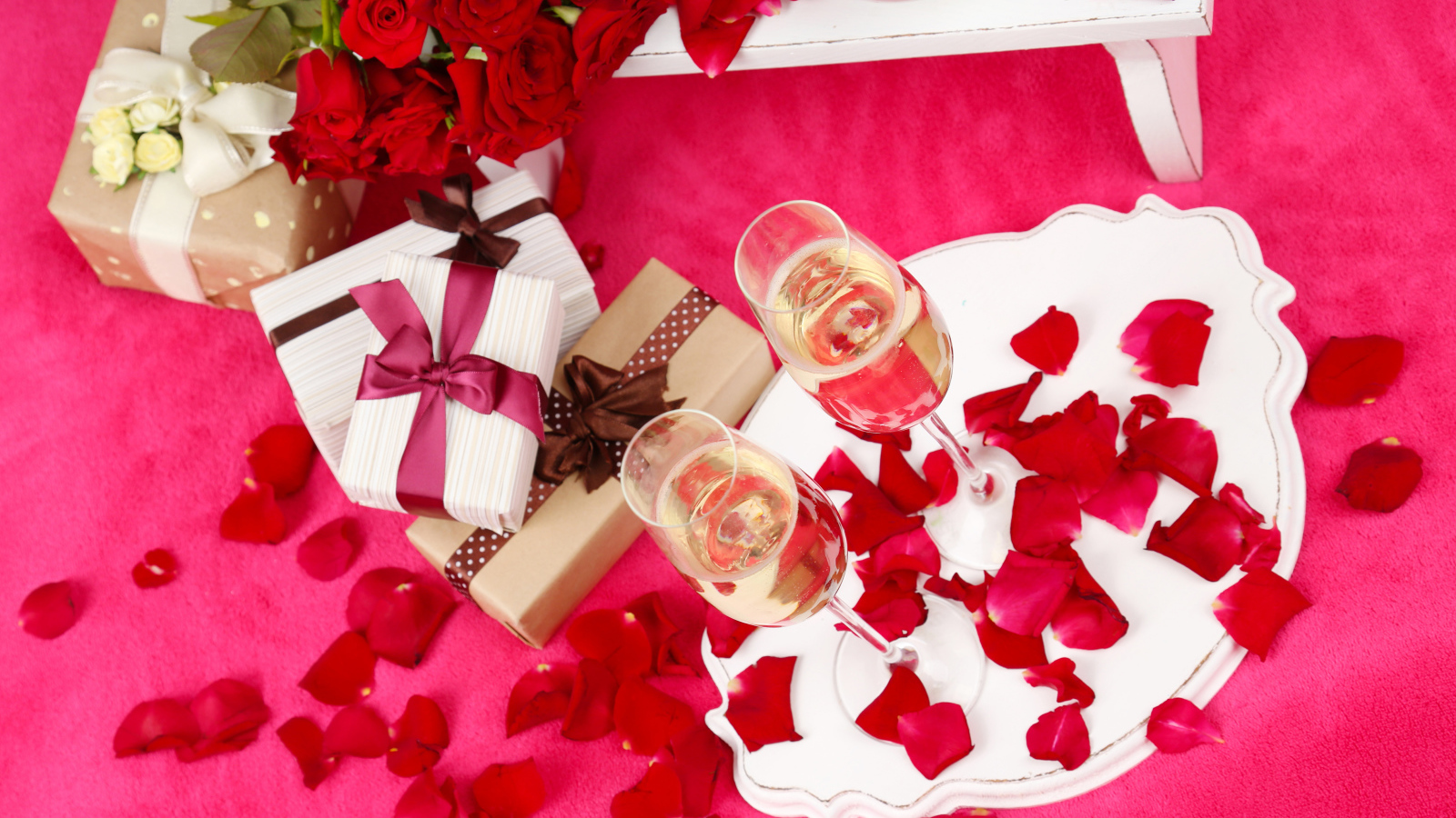 Два фужера с шампанским на красном покрывале с лепестками, подарками и букетом роз для любимой