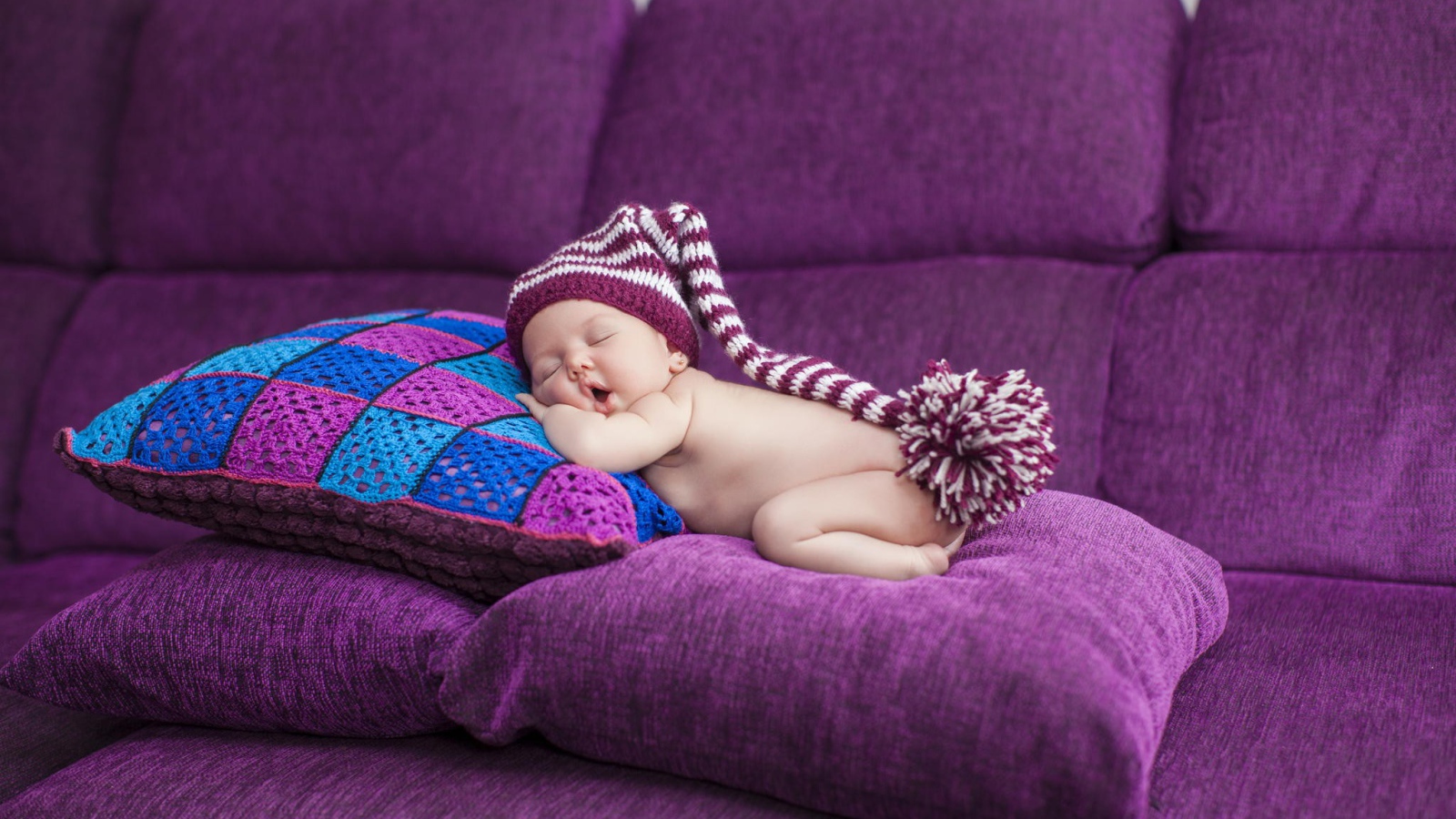 Маленький ребенок в большой шапке спит на фиолетовых подушках