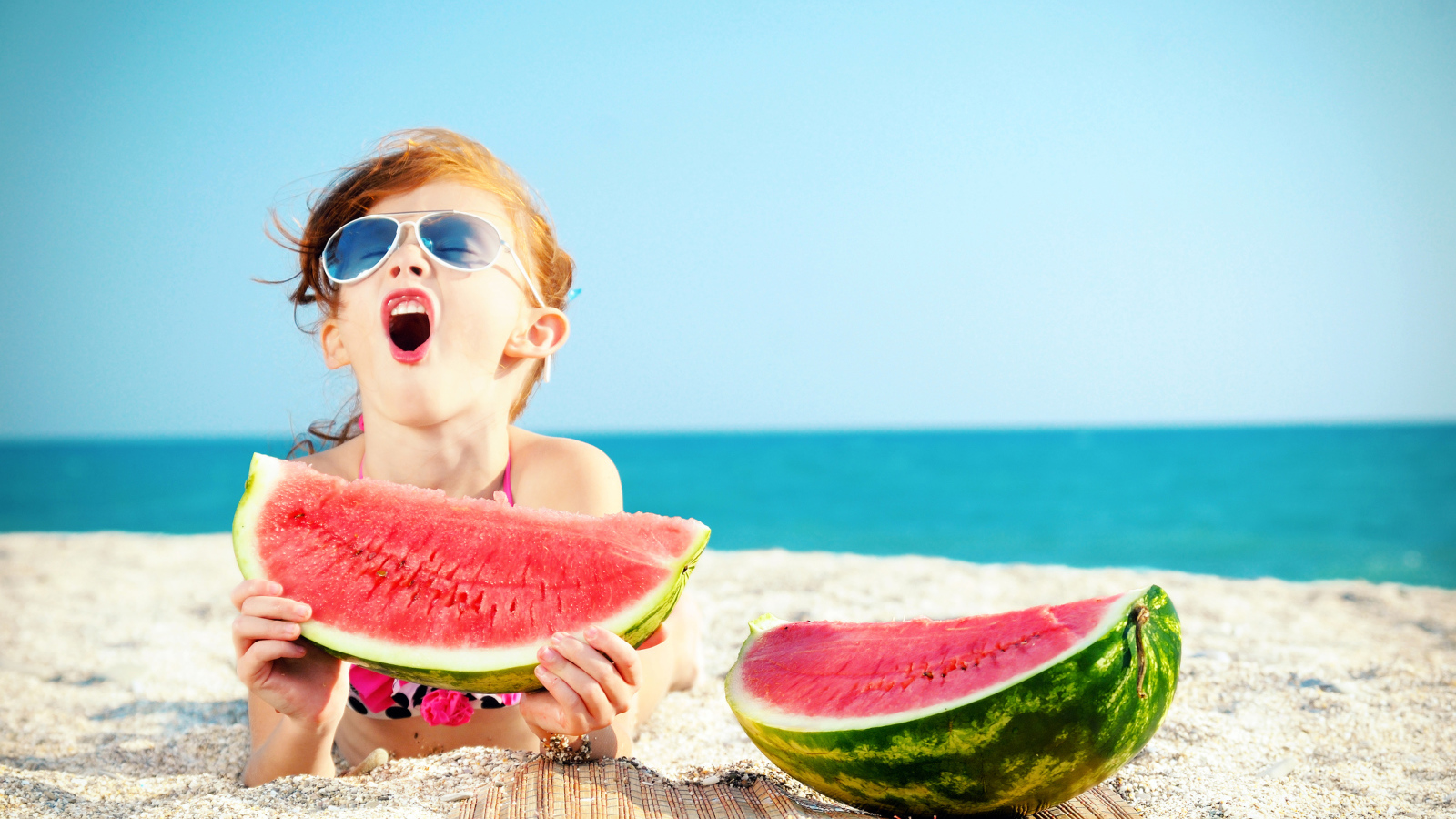 Маленькая девочка ест арбуз на песке у моря